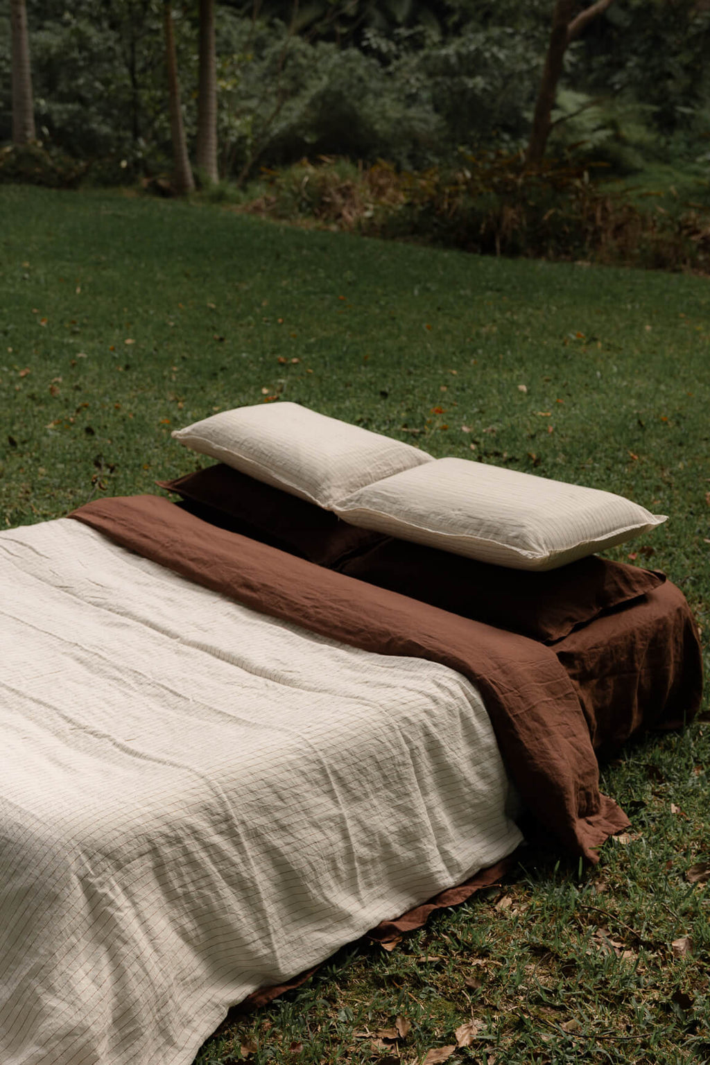 NESTD luxury linen bedding featuring Canyon Stripe white sheets and a Sable brown duvet, styled with layered pillows outdoors on the grass.