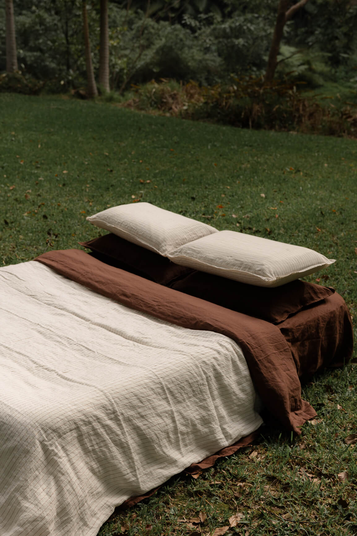 NESTD luxury linen bedding featuring Canyon Stripe white sheets and a Sable brown duvet, styled with layered pillows outdoors on the grass.