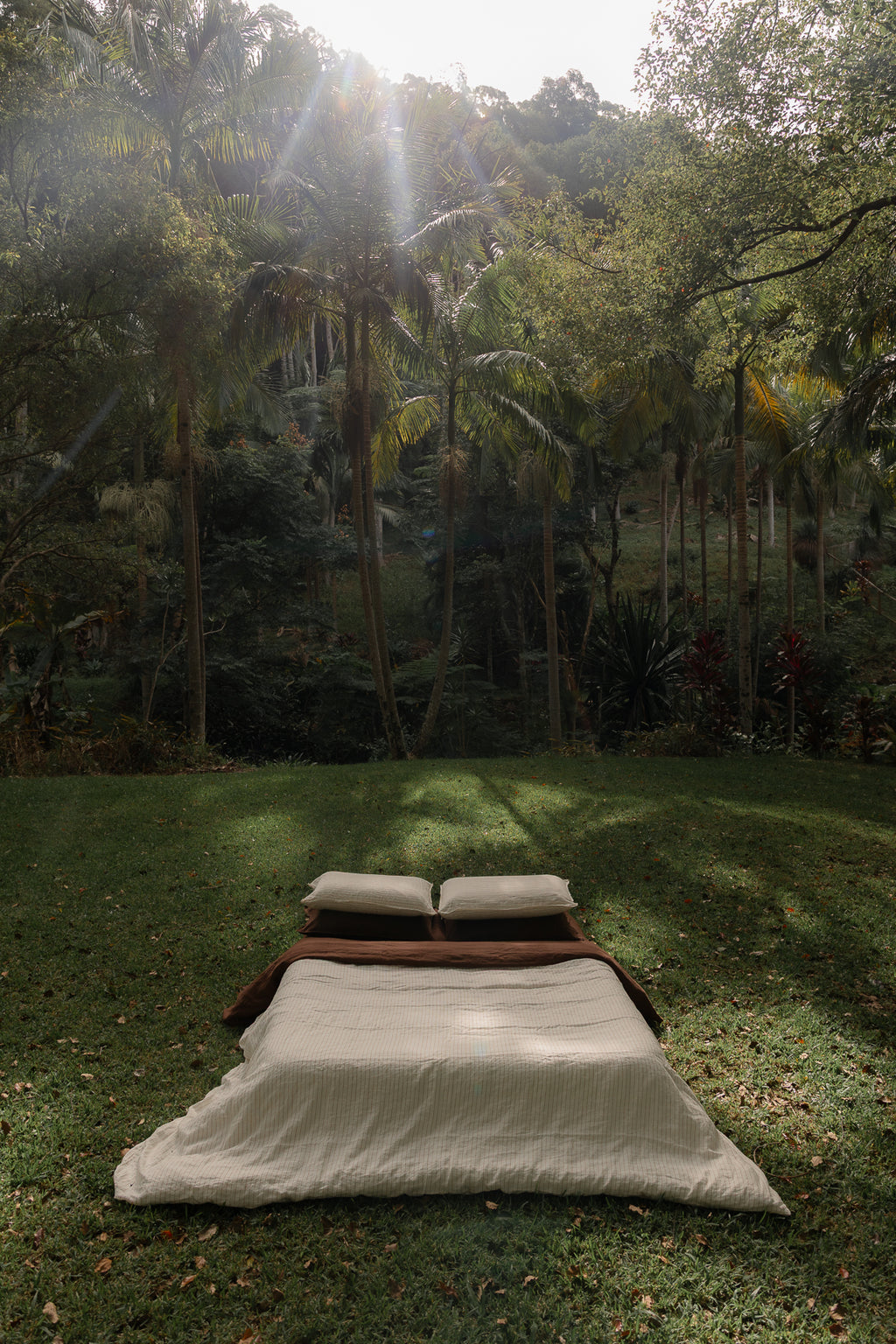 NESTD luxury linen bedding featuring Canyon Stripe white sheets and a Sable brown duvet, styled with layered pillows outdoors on the grass.
