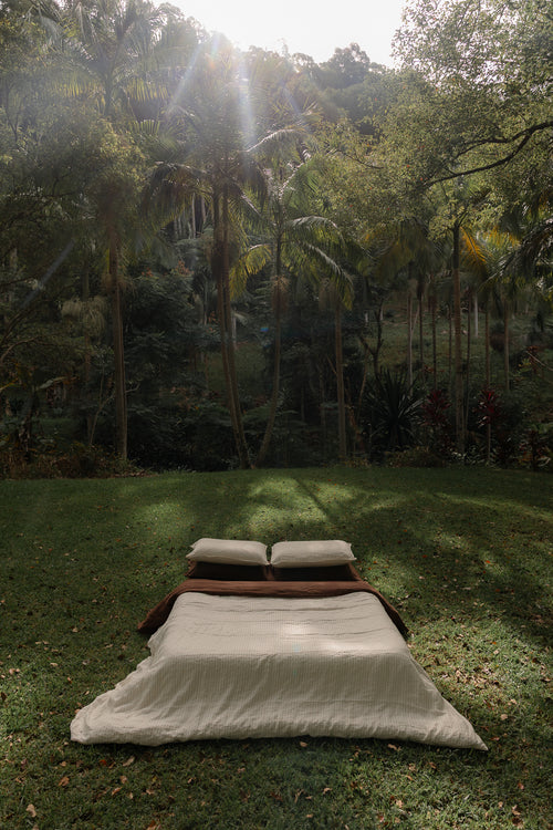 NESTD luxury linen bedding featuring Canyon Stripe white sheets and a Sable brown duvet, styled with layered pillows outdoors on the grass.
