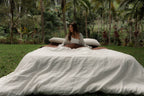 Woman sitting on NESTD luxury linen bedding featuring Canyon Stripe white sheets and a Sable brown duvet, styled with layered pillows outdoors on the grass.