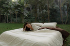 Woman lying on NESTD luxury linen bedding featuring Canyon Stripe white sheets and a Sable brown duvet, styled with layered pillows outdoors on the grass.