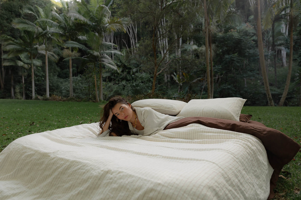 Woman lying on NESTD luxury linen bedding featuring Canyon Stripe white sheets and a Sable brown duvet, styled with layered pillows outdoors on the grass.