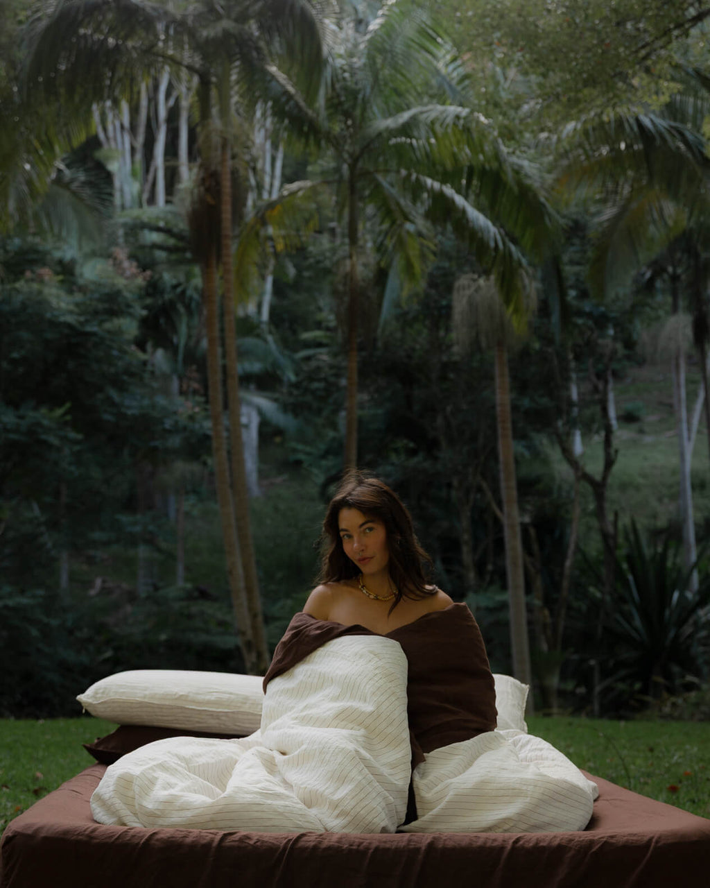 Woman wrapped in a Canyon Stripe white sheets and a Sable brown duvet, styled with layered pillows outdoors on the grass.
