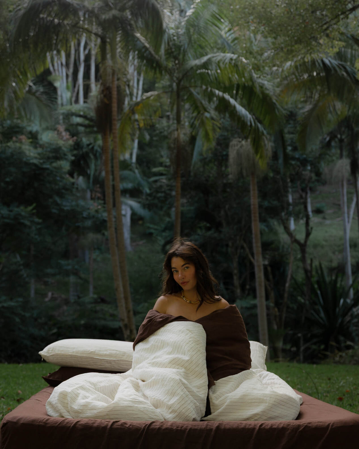 Woman wrapped in a Canyon Stripe white sheets and a Sable brown duvet, styled with layered pillows outdoors on the grass.