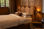 NESTD luxury flax linen bedding featuring rich Sable brown and Dune white layers, styled with matching pillows and a draped duvet against a warm textured headboard.