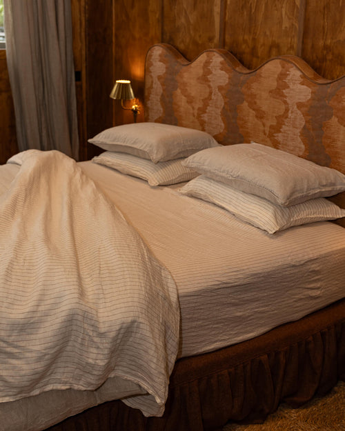NESTD luxury flax linen bedding featuring rich Sable brown and Dune white layers, styled with matching pillows and a draped duvet against a warm textured headboard.