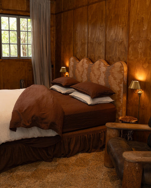 NESTD luxury flax linen bedding featuring rich Sable brown and Dune layers, styled with matching pillows and a draped duvet against a warm textured headboard.
